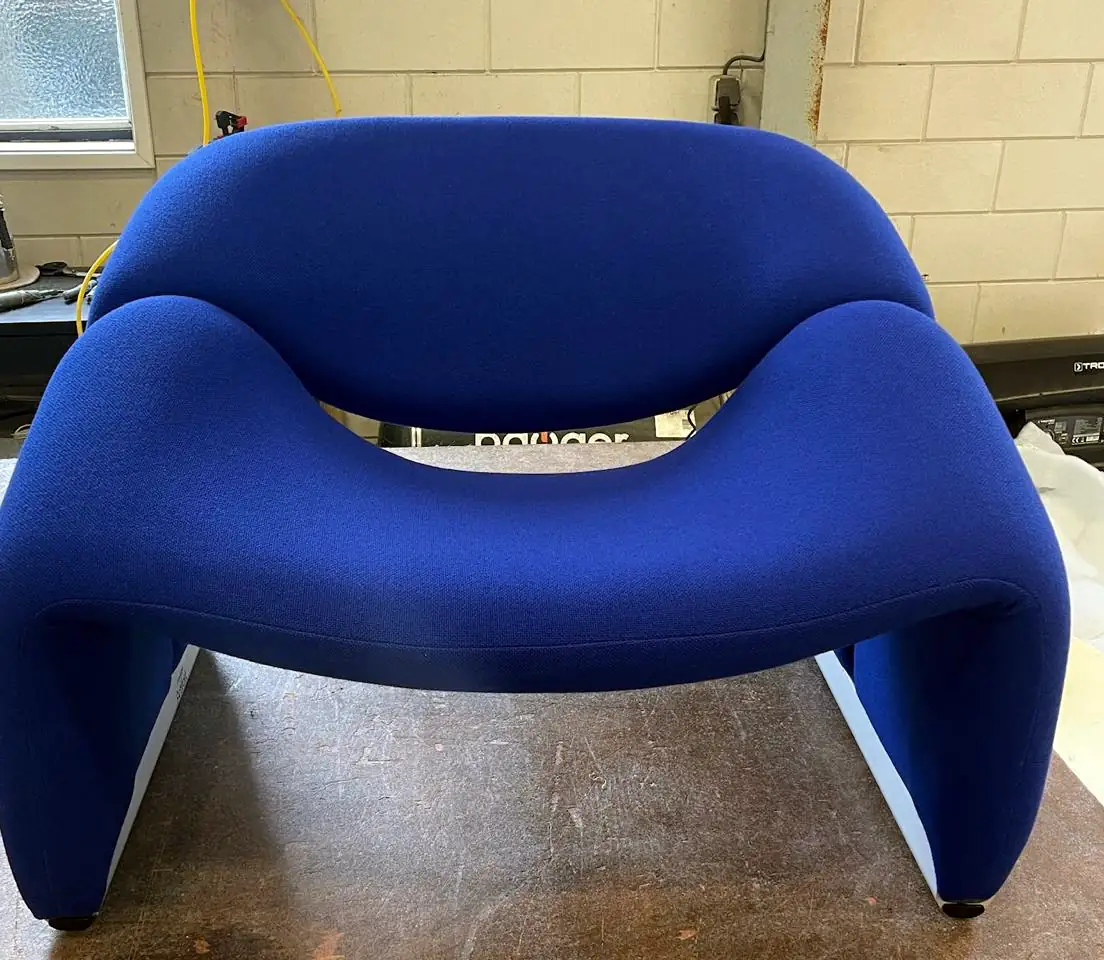 Lage designfauteuil met ronde armleuningen, opnieuw bekleed in kobaltblauwe stof, op de werkbank.